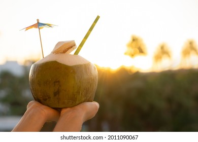  Woman hands  holding  fresh  green coconut drink with paper straw  and rainbow umbrella   on tropical  sunset background with copy space . Vacation  exotic  travel destinations  concept .