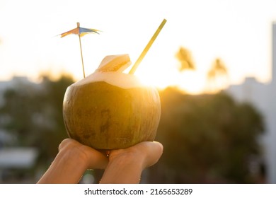  Woman Hands  Holding  Fresh  Green Coconut Drink With Paper Straw  And Rainbow Umbrella   On Tropical  Sunset Background With Copy Space . Vacation  Exotic  Travel Destinations  Concept .