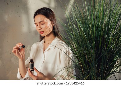 Woman Hands Holding Massage Or Cosmetics Oil Bottle For Applying Drops To Skin Of Face. Female Hold Oil On Old Wall Background With Shade From Foliage. Concept Of Healthy Lifestyle And Self Care