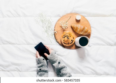 Woman Hands Holding Mobile Phone In Bed. Top View Of Breakfast In Bed