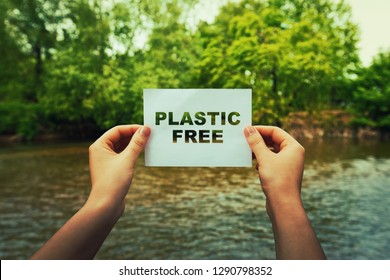 Woman hands holding a paper sheet with plastic free text in the middle of the nature near a flowing river and green forest. Ecology concept, recycle symbol, zone with no plastic.