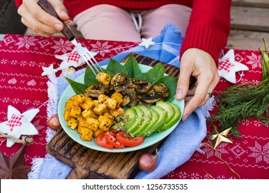 Woman Hands Holds Buddha Bowl For Christmas Lunch, Dinner. Eat Fried Tofu, Avocado Slices, Mushrooms, Tomatoes, Spinach. Take Healthy Vegetarian Food. Red Tablecloth. 