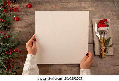 woman hands holds menu in her hands while sitting at Christmas dinner table. Christmas tree decorations border on vintage wooden table with cutlery and mockup christmas menu. - Powered by Shutterstock