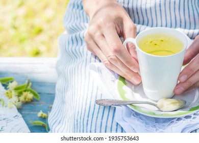 Woman Hands Keep White Cup With Fresh Linden Flowers Tea And Plate With Spoon And Honey On A Stripped Apron Background Outdoors, Person Rests On A Blue Colored Bench In The Garden