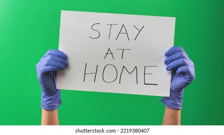 Woman Hands In Latex Blue Gloves Is Holding A Poster With Inscription Stay At Home. Doctor Arms In Medical Gloves Demonstrates Sheet With Slogan For Isolation. Close Up On Green Screen Background.