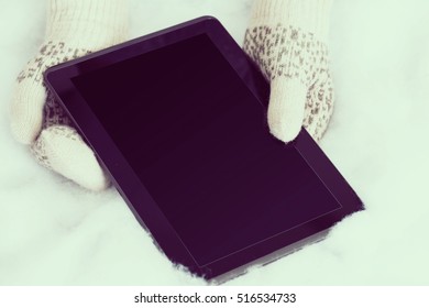 Woman Hands In Light Teal Knitted Mittens Are Holding Modern Tablet Pc On Snow Background. Winter Concept.