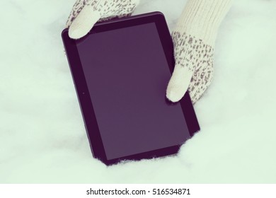 Woman Hands In Light Teal Knitted Mittens Are Holding Modern Tablet Pc On Snow Background. Winter Concept.