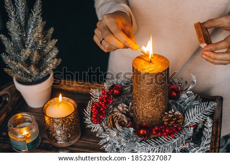 woman hands lightning big gold candle with matches in Christmas wreath. Xmas interior decoration on rustic wooden background. hygge, ornaments, candle burning in lantern on window sill at home.