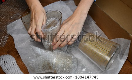 Woman hands pack up breakable fragile glassware and wrap into wrapping bubble plastic in preparation to move to new home. Packing up jug and drink glassses