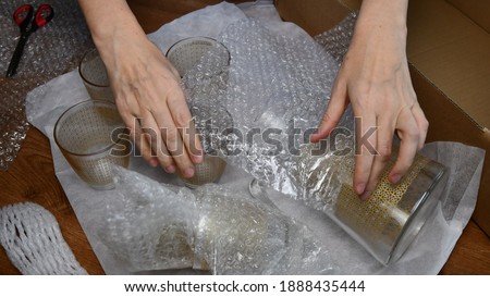 Woman hands pack up fragile glassware into wrapping bubble plastic in preparation to move in new home. Packing up jug and drink glassses. Moving day concept