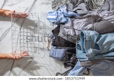 Woman hands putting stack of clean clothes for neatly tidying up and placing in steel wire wardrobe baskets organiser on the white marble table. Marie Kondo style and space saving storage concept.