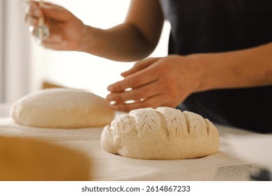 Las manos de la mujer marcan hábilmente la superficie de un pan de masa redonda, preparando pan de masa madre para hornear. El luminoso entorno de la cocina ofrece un ambiente cálido e ingredientes frescos.