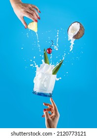Woman hands support fly glass of tasty Frozen Pina Colada Traditional Caribbean cocktail with splash,  ingredients falling in glass. Blue background