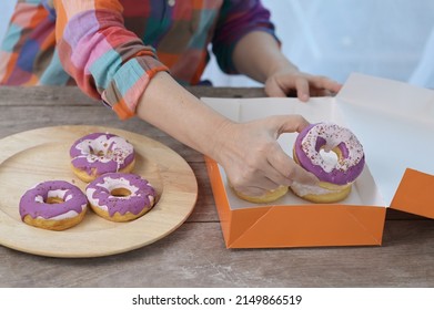 Woman hands is taking purple colored glazed donuts in ornage color donut box and arranged in a large wooden tray. Asian women in bright colored shirts arrange donuts (Unhealthy snack) on wooden trays.