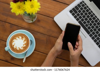 Woman Hands Using Mobile Phone And Cup Of Coffee Have A Abstract Milk Froth Put On Wooden Table And Have Brew Spoon Put Near A Cup,have A Blurry Yellow Flower And Notebook In Top View Coffee Concept