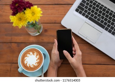 Woman Hands Using Mobile Phone And Cup Of Coffee Have A Abstract Milk Froth Put On Wooden Table And Have Brew Spoon Put Near A Cup,have A Blurry Yellow Flower And Notebook In Top View Coffee Concept
