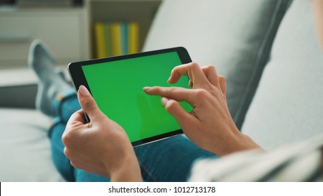 Woman hands using tablet with green screen, lieng on sofa at home. View from the back. Chroma key.