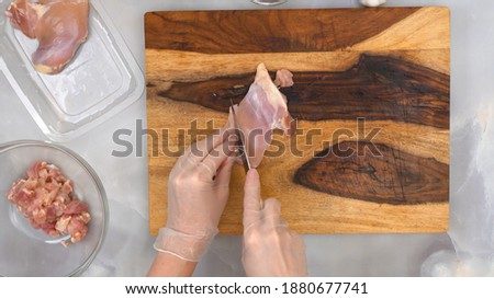 Woman hands wearing gloves cut chicken fillet on wooden cutting board, top view