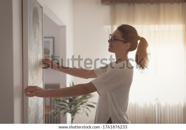 Woman hanging a painting at home and decorating her\
contemporary living room