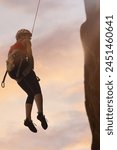 A woman is hanging from a rope while wearing a red tank top. The sky is pink and the sun is setting