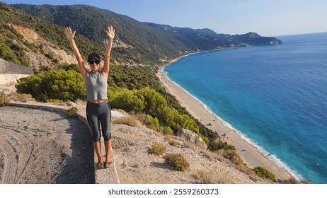 Woman happily posing with arms raised on scenic coastal cliff overlooking turquoise waters, sandy beach and mountains, a perfect summer vacation destination. panoramic view towards Pefkoulia beach - Powered by Shutterstock