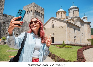 A woman happily takes a selfie photo of Manasija Monastery in Serbia, using her smartphone to document its historic architecture - Powered by Shutterstock