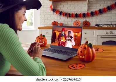 Woman Happy To See Her Friends At Virtual Halloween Costume Party. Smiling Witch Sitting In Front Of Laptop Computer At Kitchen Table Decorated With Pumpkins And Having Online Celebration With