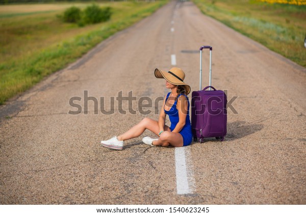 A\
woman in a hat and with a big bag sits on the pavement. Girl in a\
blue tight-fitting dress with a purple suitcase on the roadway. A\
brunette with a sports figure is waiting for a\
bus.