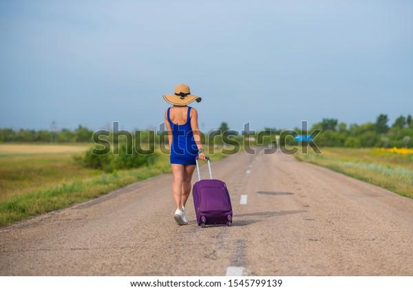 A woman\
in a hat and with a big bag is walking along the roadway. Girl in a\
blue tight-fitting dress with a purple suitcase on the track.\
Brunette with a sports figure on the\
road.