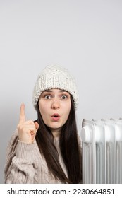 A woman in a hat getting warm near the oil heater and looks and points up with her finger.