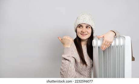 A woman in a hat hugs an oil heater and points her finger to the side in place for text.