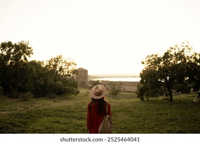 Woman in hat and orange jacket stands in a field looking at an ancient fortress during sunset, surrounded by nature, enjoying peaceful moments while traveling. - Powered by Shutterstock