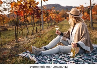 Woman with hat and poncho drinking white wine. Female vintner sitting on blanket in vineyard and relaxing at fall season. Relaxation in autumn winery - Powered by Shutterstock