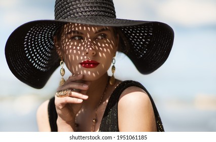 Woman In Hat Portrait. Fashion Luxury Model In Black Summer Hat With Make Up And Golden Jewelry. Close Up Beauty Face Over Sky Background