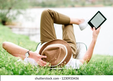 Woman with hat reads e book from kindle device while lying in the grass near the lake.