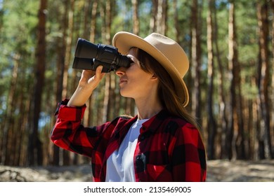 Woman In Hat And Red Plaid Shirt Looking Through Binoculars In The Forest.