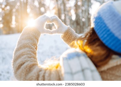 A woman in a hat and scarf against the backdrop of a snowy winter forest holds her hands in the shape of a heart. Back view. Young woman enjoying a sunny winter day. Vacation concept, lifestyle. - Powered by Shutterstock