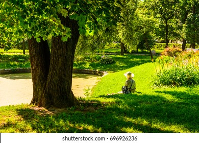 Woman In A Hat Sits On The Shore Of A Pond In The Park On A Sunny Morning In The Shade Of A Tree