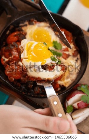 Woman is having breakfast on summer terrace: israeli shakshuka with tomatoes, salami, chili pepper and fried eggs, served in hot cast pan with toasts