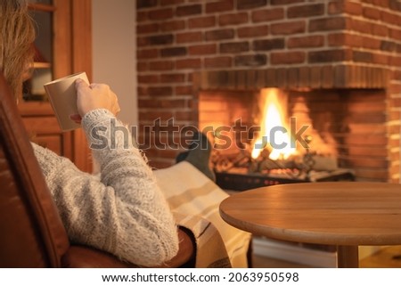Woman having a cup of coffee in front of the fire of a fireplace in winter.