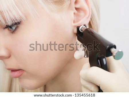 Woman having ear piercing process with special equipment in beauty center by medical worker.