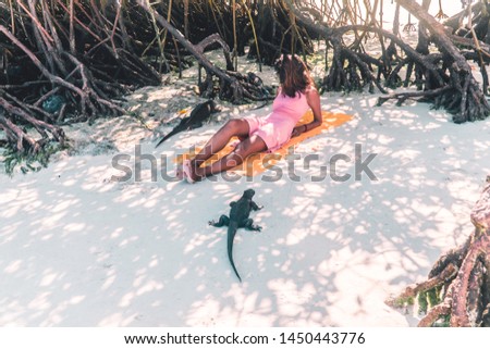Woman having fun, sunbathing with an IGUANA reptile. Beautiful girl in pink sitting on beach sand. Holiday, vacation vibes. Asian Latino girl. Shot in San Cristobal, Galapagos Islands.
