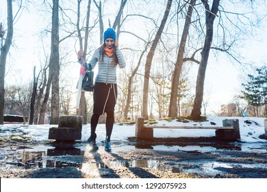 Woman Having To Step Into A Puddle Of Water And Thawing Snow, While Speaking On The Phone In The Park