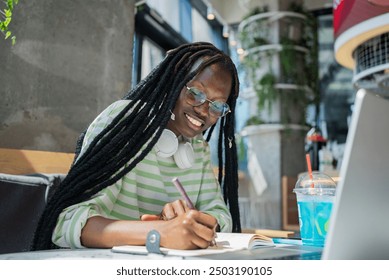 Woman with headphones and glasses smiles while studying and writing in a cafe, enjoying a productive and relaxed environment. African-American student is finishing her term paper at university cafe.  - Powered by Shutterstock