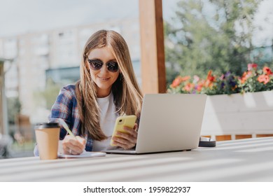 Woman In Headphones Having A Video Call Chat With Cellphone. Happy And Smiling Girl Working Outside And Drinking Coffee. Using Computer And Mobile Phone. Distance Learning Online Education And Work.