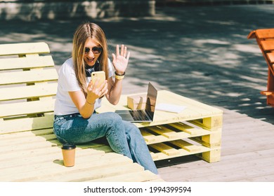 Woman In Headphones Having A Video Call Chat With Cellphone. Happy And Smiling Girl Working Outside And Drinking Coffee. Using Computer And Mobile Phone. Distance Learning Online Education And Work.