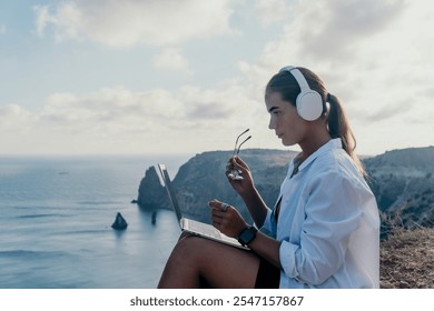 Woman Headphones Laptop Cliffside Ocean View - Relaxing and working while enjoying the beautiful seascape. - Powered by Shutterstock