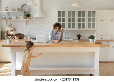 Woman In Headphones Working Or Studying Online At Home While Little Daughter Running Around Playing Active Game, Young Mom Freelancer Sitting At Kitchen Table With Laptop, Writing Taking Notes