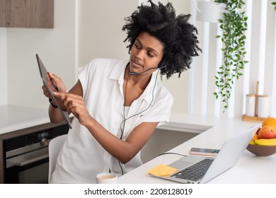 Woman With Headset Sitting At Computer Desk, Looking At Tablet. Serious Female Employee Working From Home, Having Online Video Call. Home Office, Communication, Connection, Technology Concept