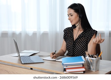 Woman in headset taking notes while passing online test via laptop at table indoors
